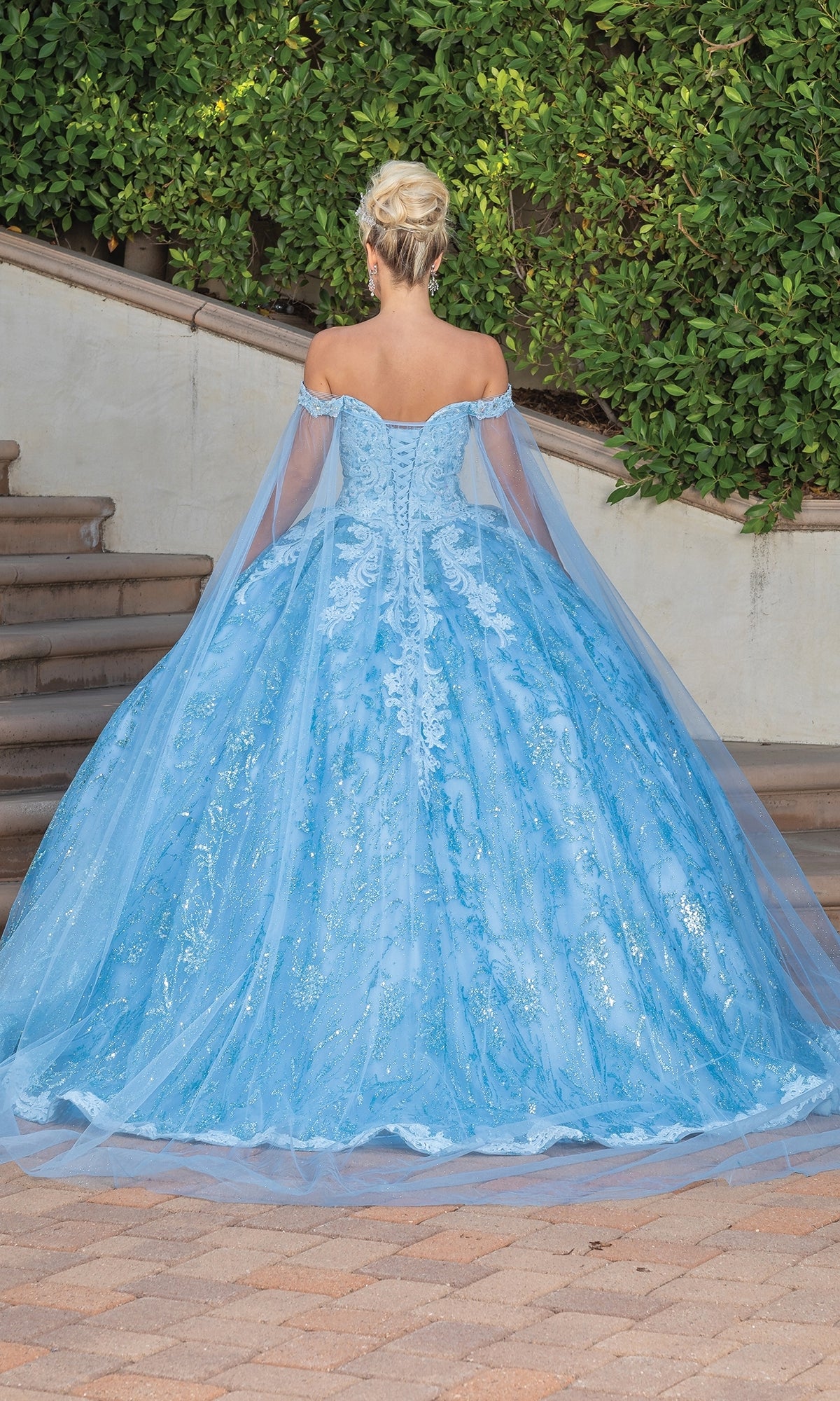 A woman with blonde hair in an updo wears the Quinceanera Dress 1782 By Dancing Queen, a light blue off-the-shoulder gown with lace, tulle details, and a sweetheart neckline, standing outdoors with her back to the camera.